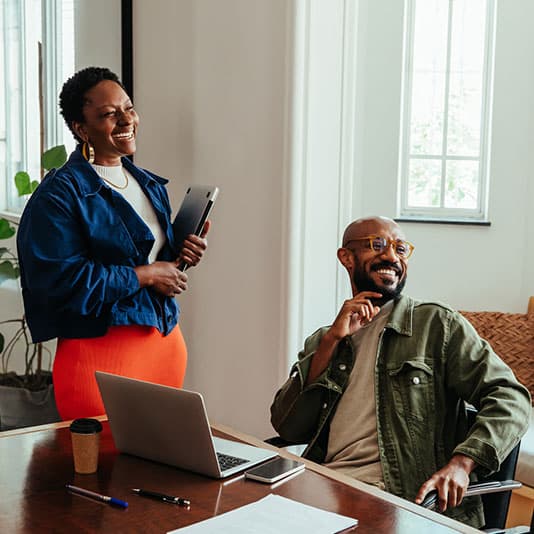 Two people in a bright office, one standing with a tablet, the other seated at a desk with a laptop, both smiling and engaged.