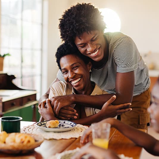 A son hugs his mother from behind at a dining table, both smiling. Plates of food and drinks are on the table. Bright, cheerful atmosphere