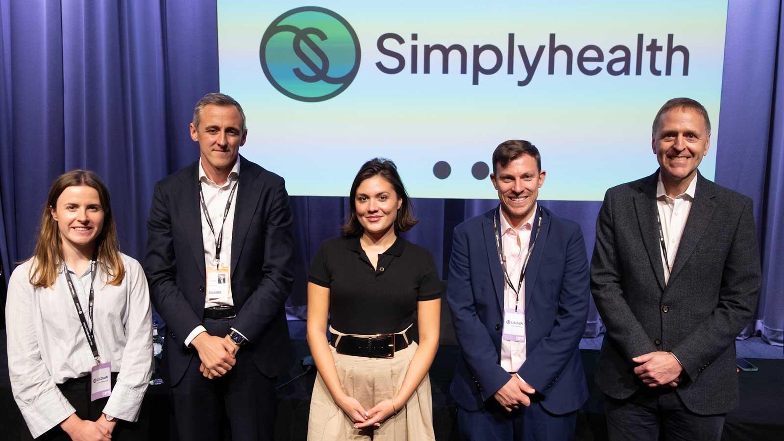 Five people standing in front of a "Simplyhealth" sign, smiling, dressed in business attire
