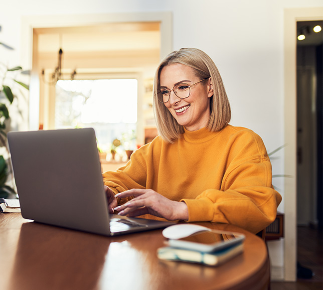 Smiling woman in a yellow sweater typing on a laptop at a wooden table, with a smartphone and mouse nearby, in a bright room.