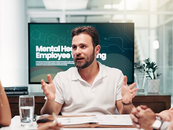 Man in a white shirt speaking at a meeting, with "Mental Health Employee Training" displayed on a screen in the background.