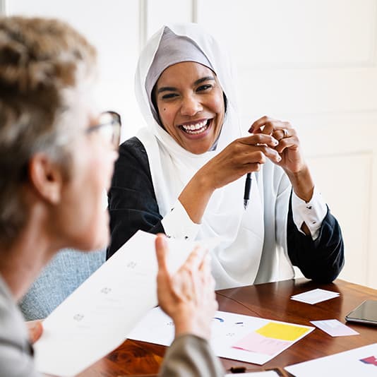 Two women smiling and talking at a table, one wearing a hijab.