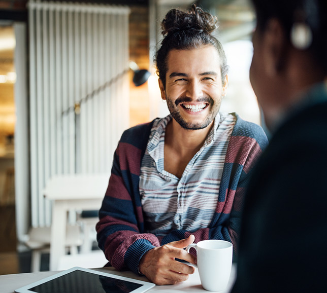 Man sat with coffee mug and tablet device, smiling with friend